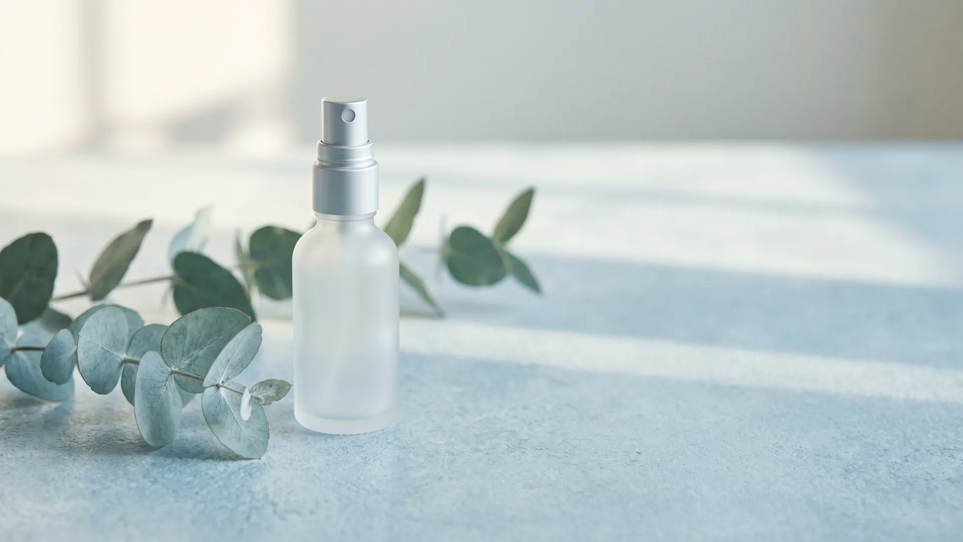 A small unlabeled frosted spray bottle beside eucalyptus on a pale blue stone surface