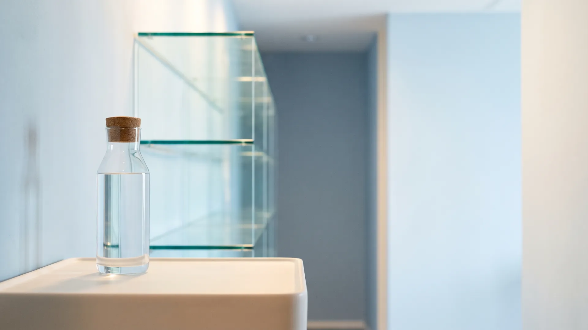 A corked glass carafe of water in front of empty glass shelves in a pale blue room
